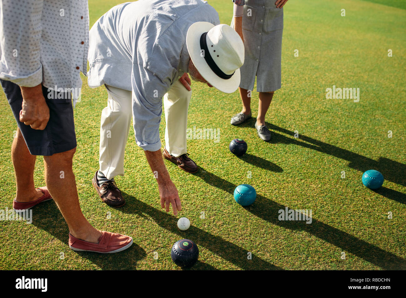 Senior uomo in hat la piegatura verso il basso per scegliere un Legname in blocco in un prato. Uomo anziano giocando a bocce in un prato con i suoi amici. Foto Stock