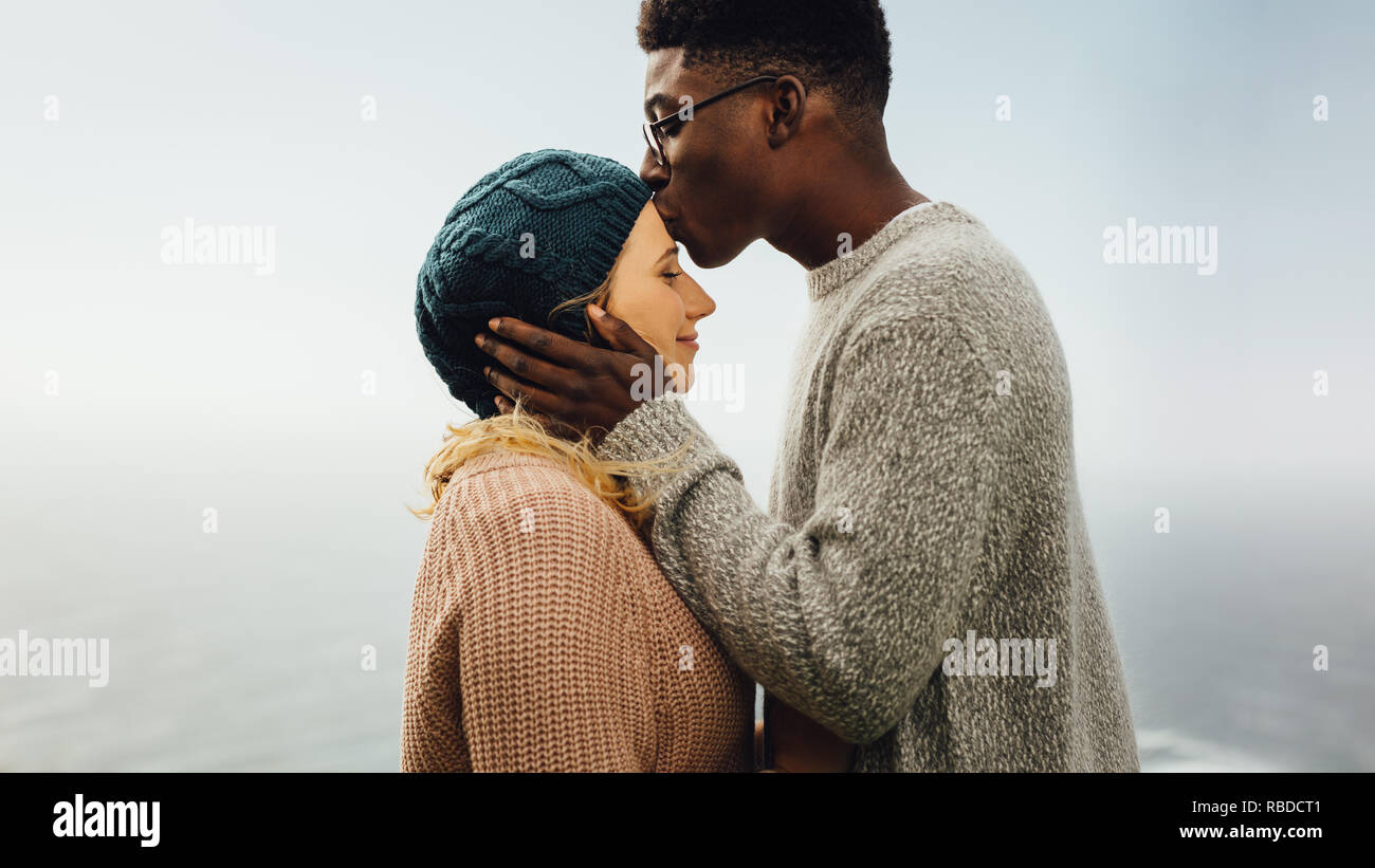 Vista laterale di amare interracial coppia insieme all'esterno. Giovane uomo baciando la fronte della sua fidanzata. Foto Stock