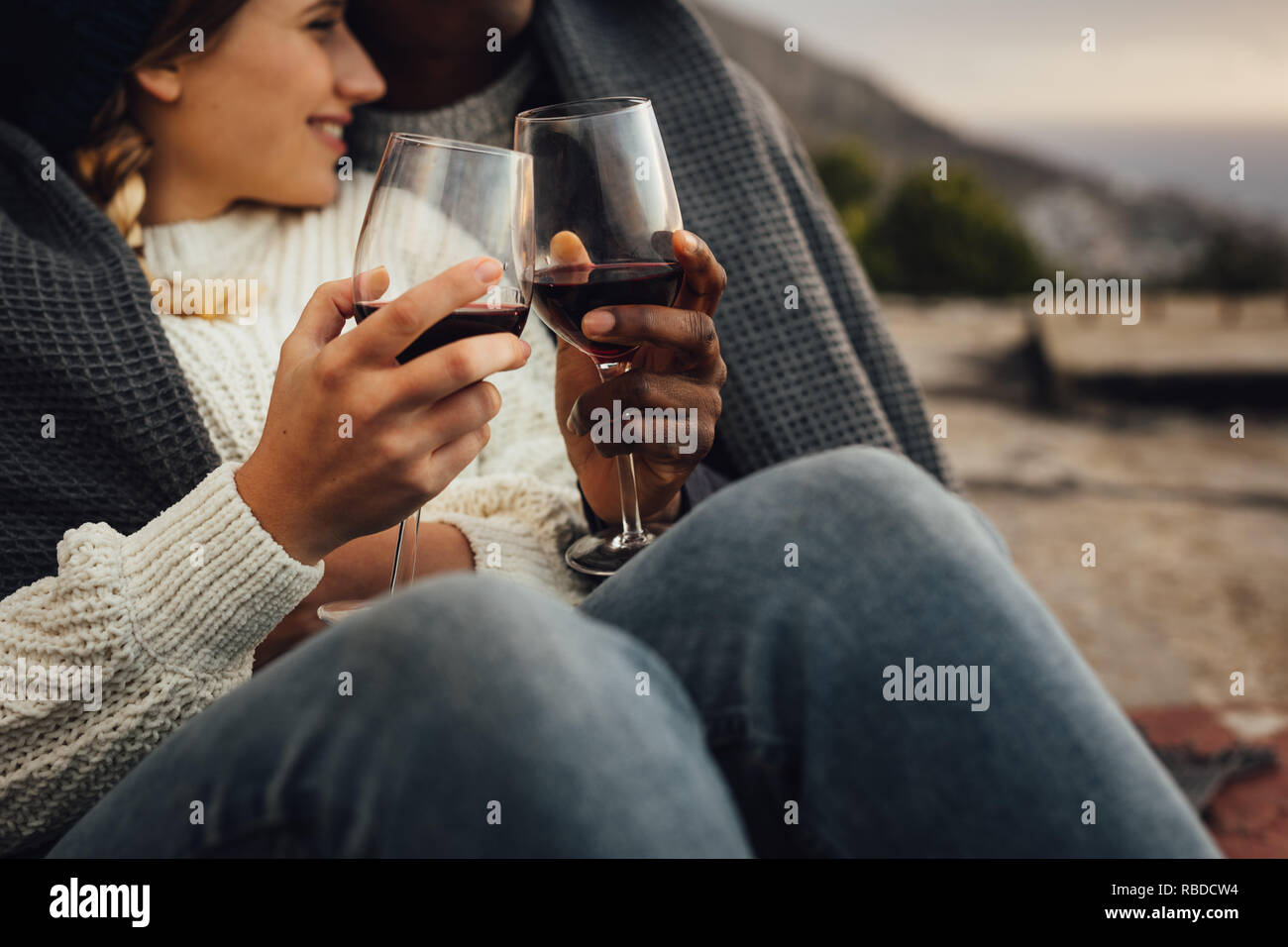 Ritagliato shot di un uomo e di una donna avvolta in una coperta seduti insieme vino di tostatura durante un picnic. Giovane godendo un picnic all'aperto con vino su un Foto Stock