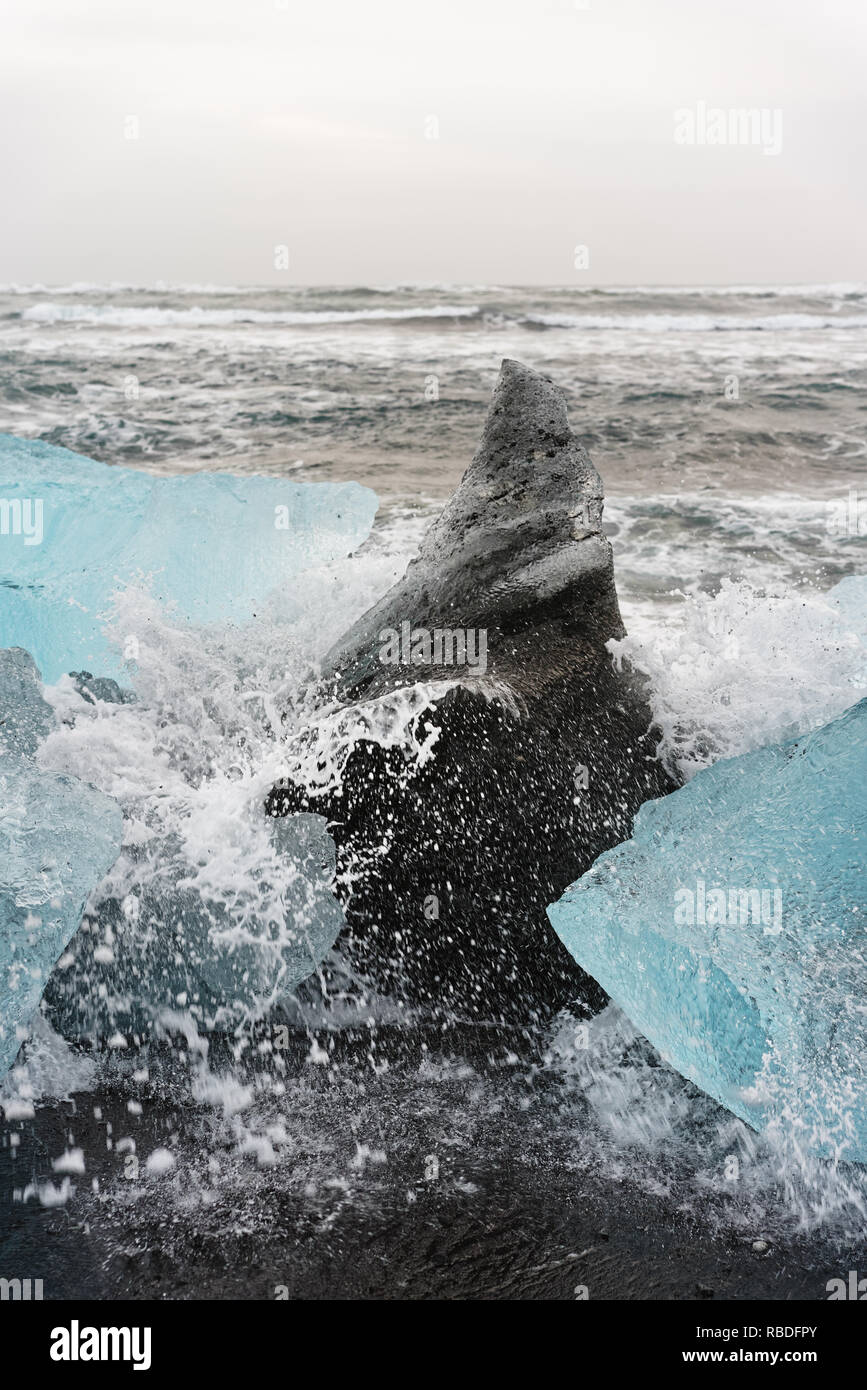 Blocchi di ghiaccio in blu e sfumature scure su una spiaggia nera con forte surf, spray di una onda schizzi sul ghiaccio, onde roll in background - Posizione: Foto Stock