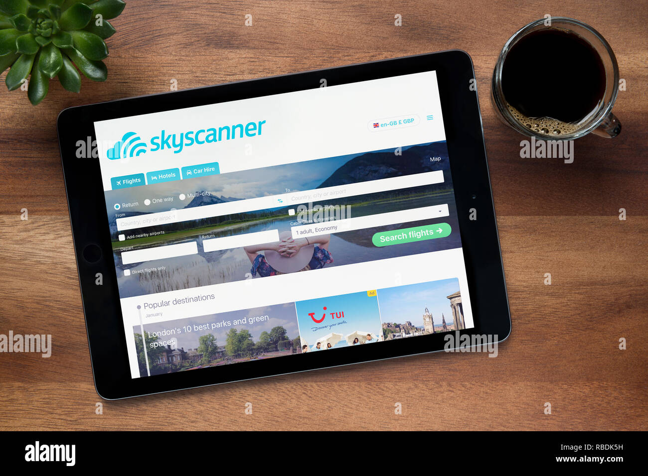 Il sito web di Skyscanner è visto su un tablet iPad, su di un tavolo di legno lungo con un caffè espresso e una pianta di casa (solo uso editoriale). Foto Stock