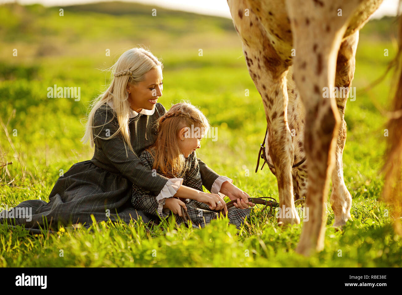 Vista laterale della giovane madre con bambina in abiti abbracciando a macchie di cavalli sul prato verde Foto Stock