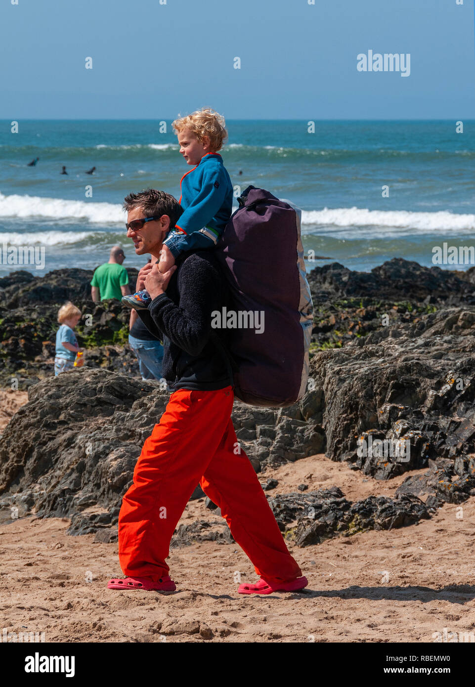 Padre camminando lungo la spiaggia in una giornata di sole e dando il suo bimbo figlio un piggyback ride su Croyde Beach, Croyde, Devon, Regno Unito Foto Stock