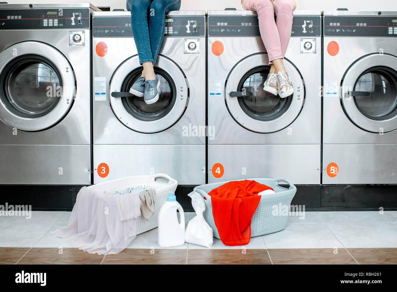 Donna seduta su macchine di lavaggio in attesa di lavaggio al servizio di lavanderia self-service con ceste piene di vestiti. Immagine ritagliata con nessun volto Foto Stock
