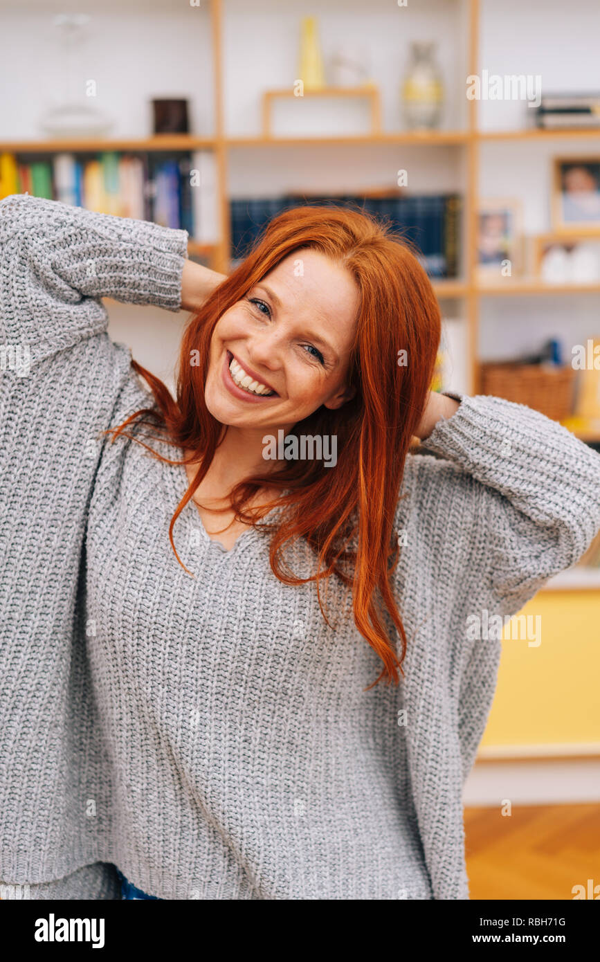 I giovani i capelli rossi donna in grigio maglione guardando la fotocamera con bel sorriso, tenendo le mani dietro la nuca. Parte anteriore a metà lunghezza ritratto in interni Foto Stock