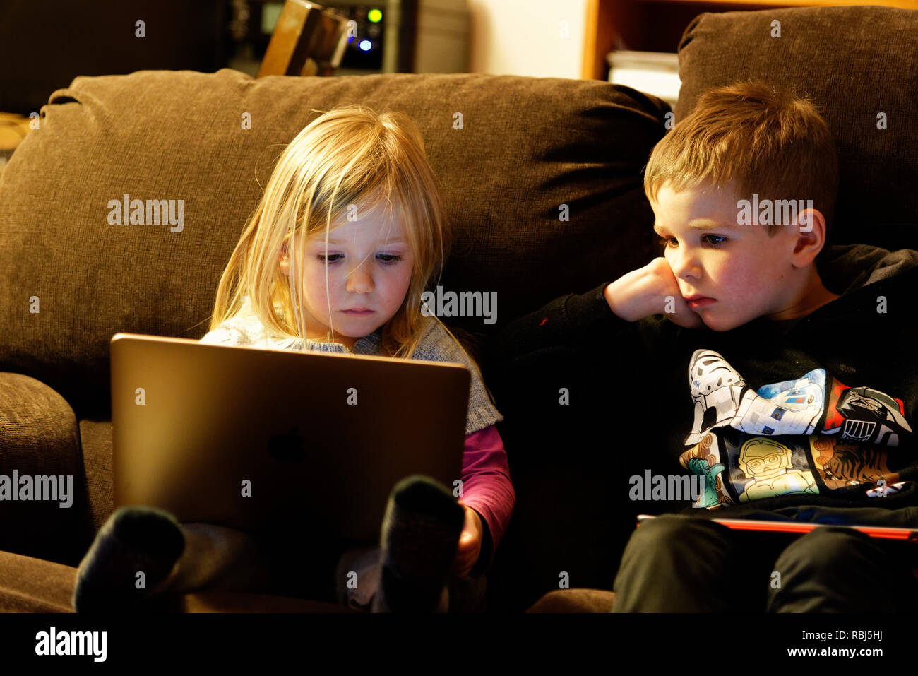 Una bambina di 4 anni seduto su un divano con un computer portatile mentre suo fratello (6 anni) orologi. Foto Stock