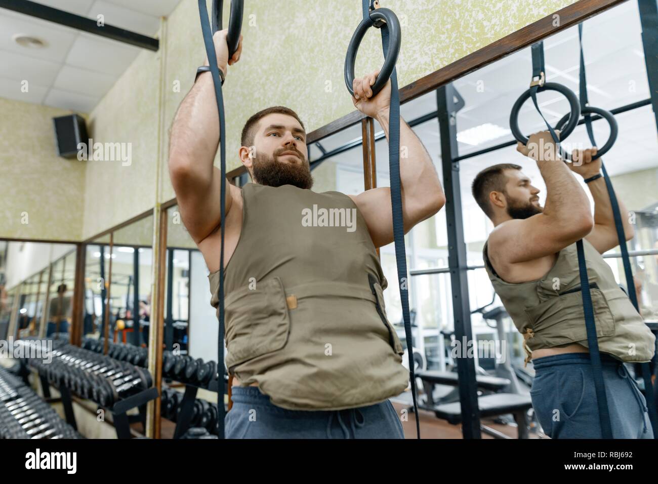Muscoloso caucasica uomo barbuto facendo esercizi vestito con giubbotto ponderata in palestra, in stile militare. Foto Stock