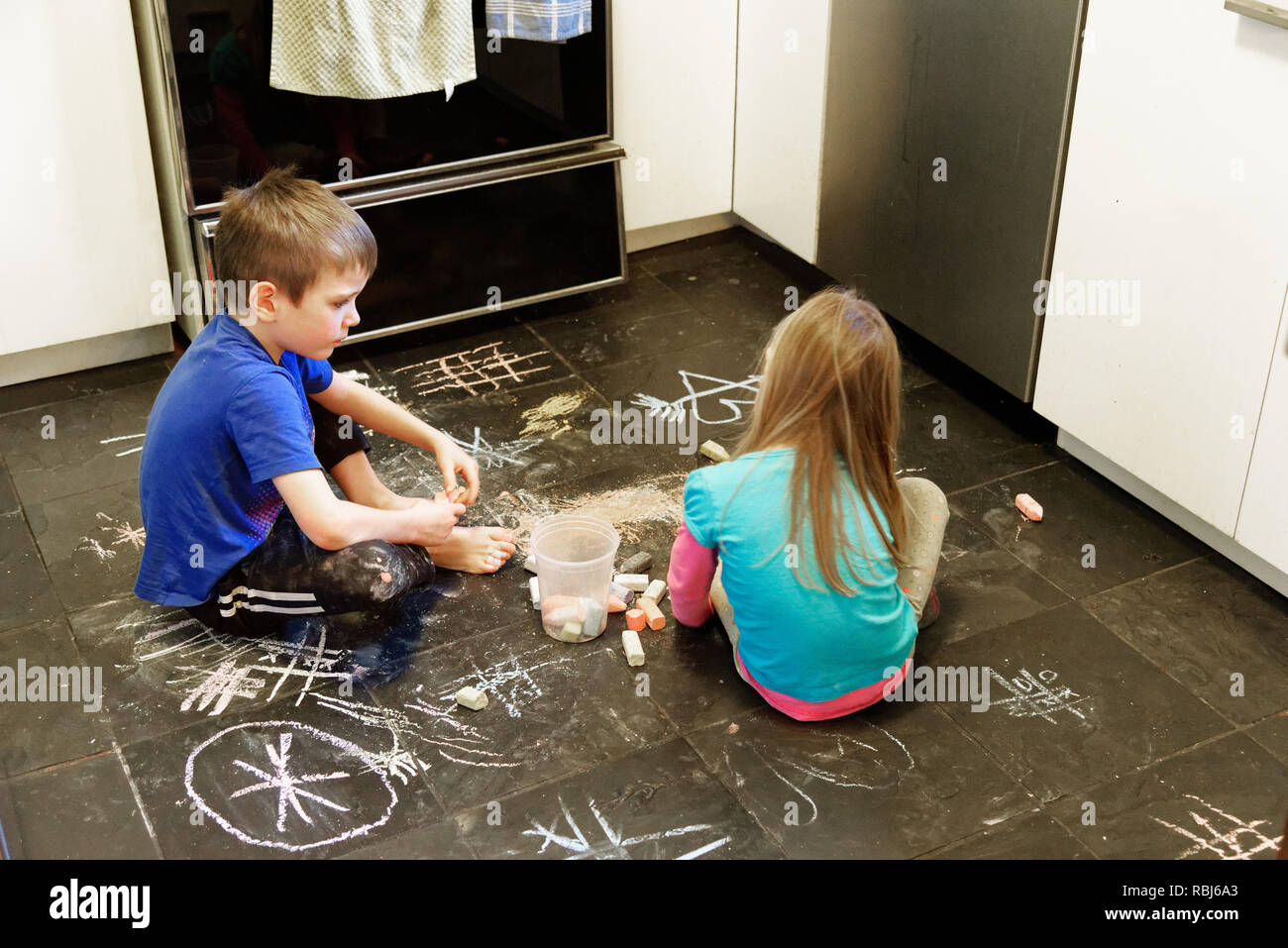 Due bambini che utilizzano i gessi per disegnare su un pavimento di ardesia nella loro cucina Foto Stock