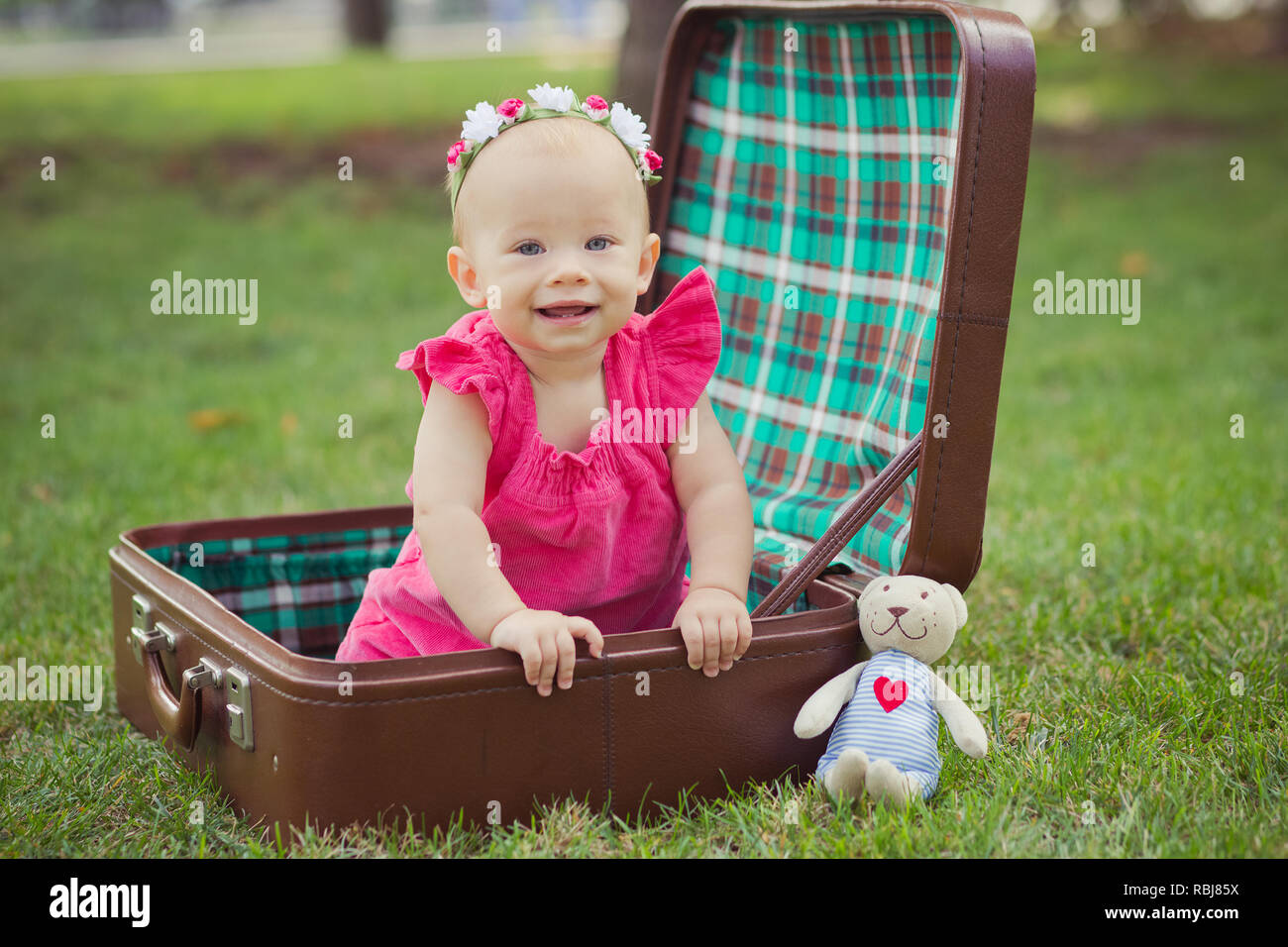 Carino baby ragazza bionda bambino indossando abiti rosa seduta nel verde parco natura su marrone old fashion retrò borsa trunk con Teddy bear amico. Foto Stock