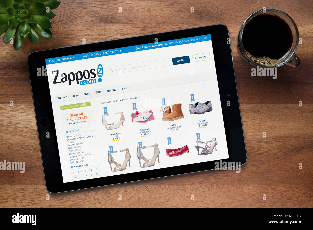 Il sito web di Zappos è visto su un tablet iPad, su di un tavolo di legno lungo con un caffè espresso e una pianta di casa (solo uso editoriale). Foto Stock