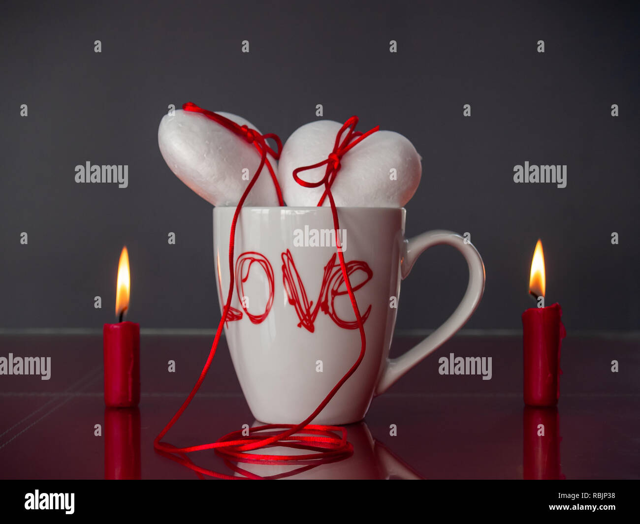 Concetto di amore poliespan due cuori uniti con un filo rosso che simboleggia la leggenda del filo rosso su una tazza colazione e due rosso acceso candele Foto Stock