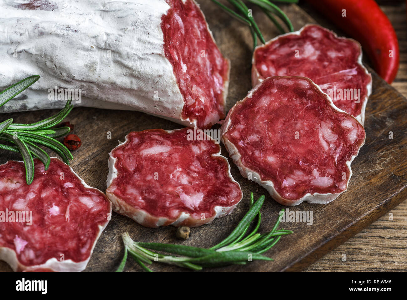 Salame francese con rosmarino, peperoncino e pepe in grani sul tagliere di legno su sfondo rustico. close up Foto Stock