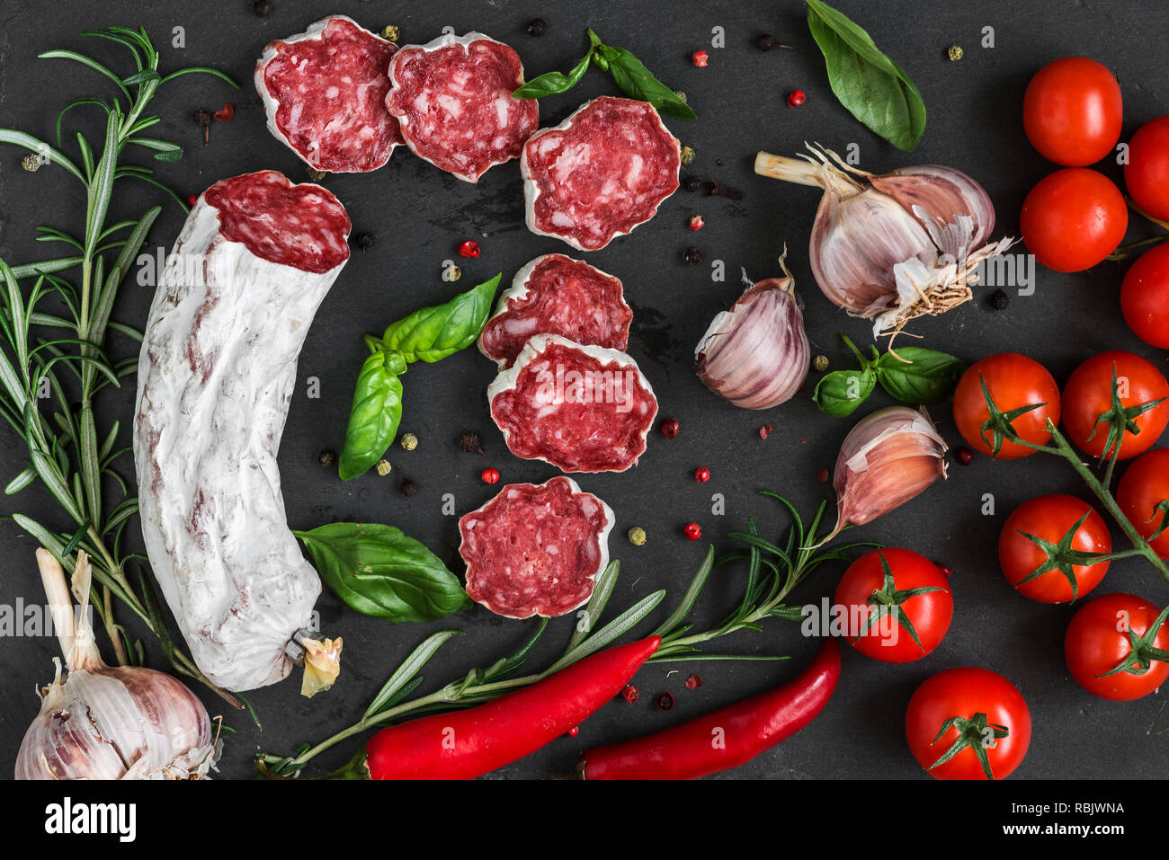 Fette di salame con spezie, peperoncino, pomodoro, aglio e rosmarino nero ardesia sfondo. vista superiore. laici piatta Foto Stock
