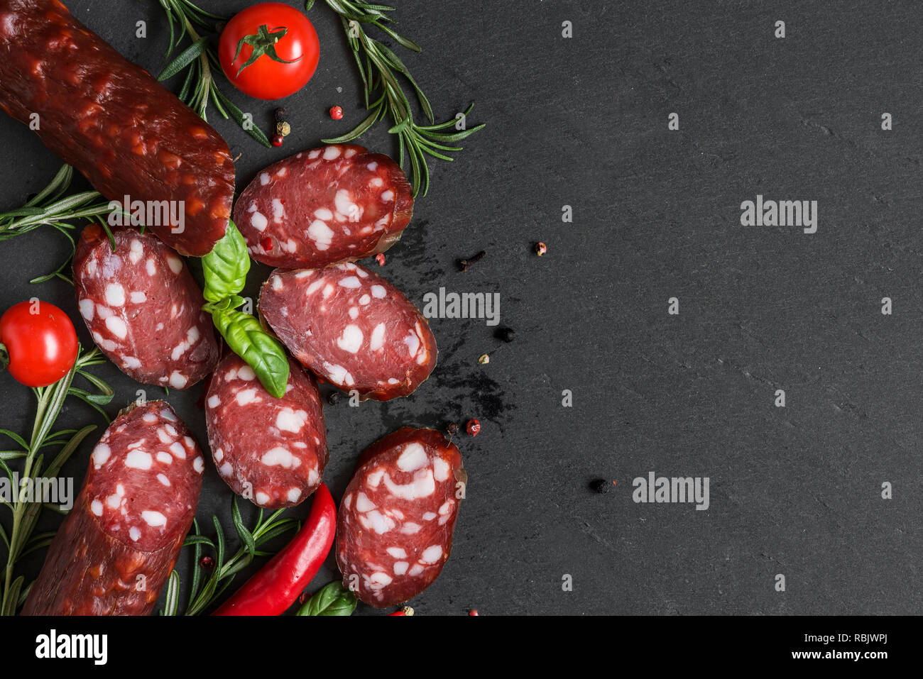 Fette di salame con spezie, pomodori e rosmarino nero ardesia sfondo. vista superiore con spazio di copia Foto Stock
