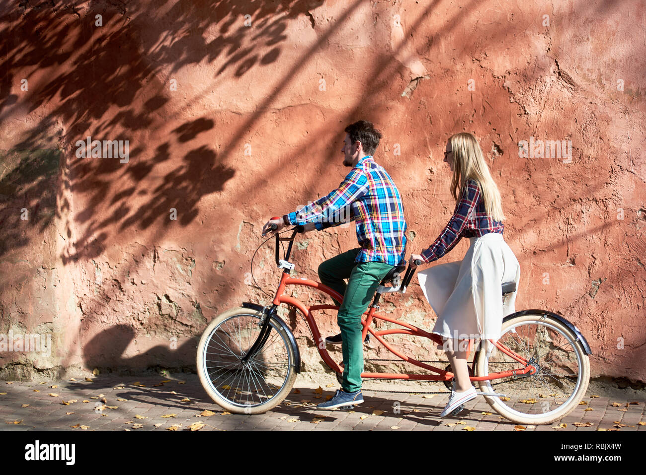 Giovane turista attraente giovane in abbigliamento casual, barbuto uomo e donna bionda in bicchieri di ciclismo biciclette tandem lungo vuoto pavimentato soleggiata sul marciapiede Foto Stock