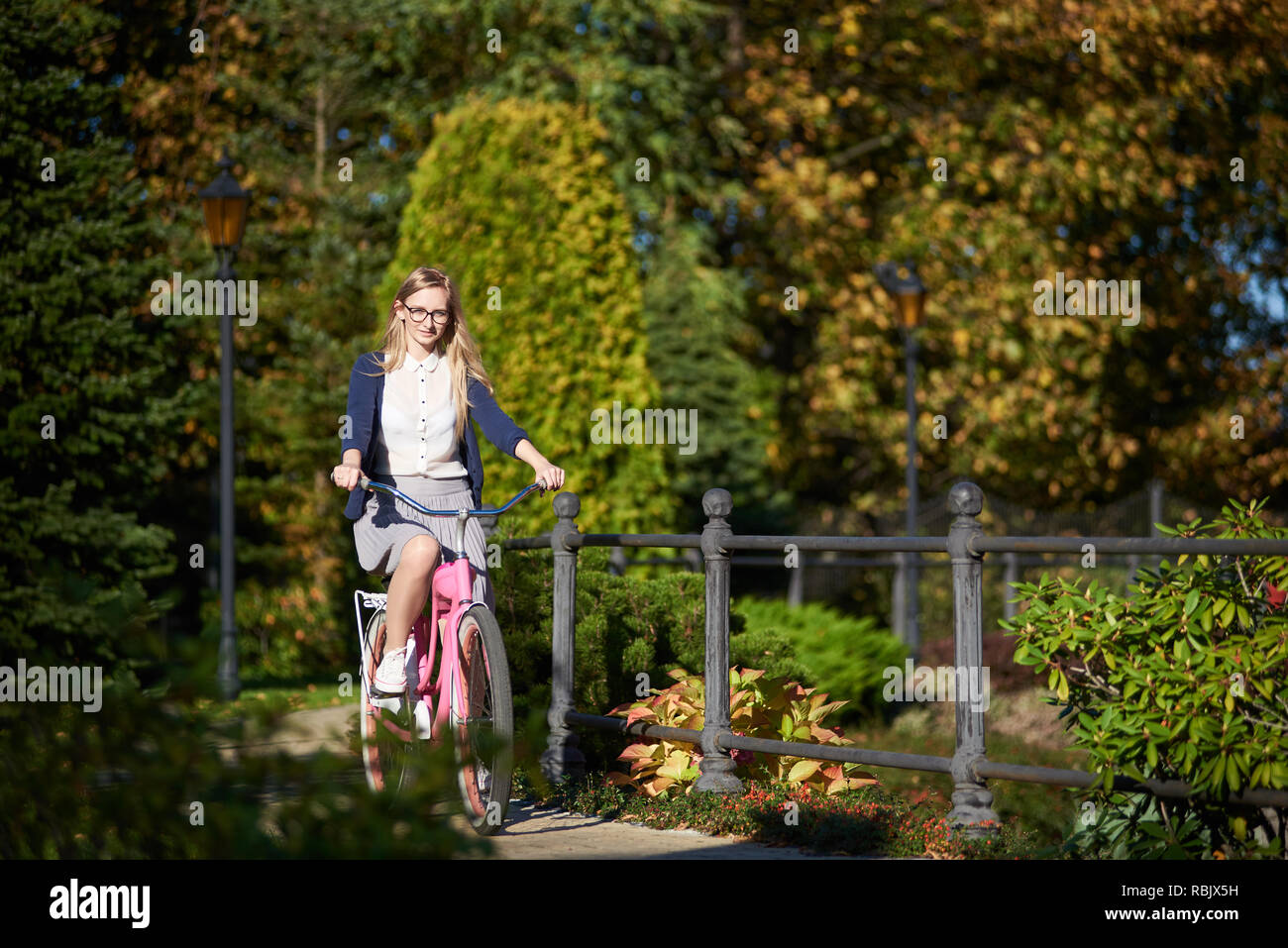 Felice bionda con i capelli lunghi attraente traveler donna in bicchieri e abbigliamento casual ciclismo moderno pink lady bike lungo stretto ponte di parco sul bokeh backg Foto Stock