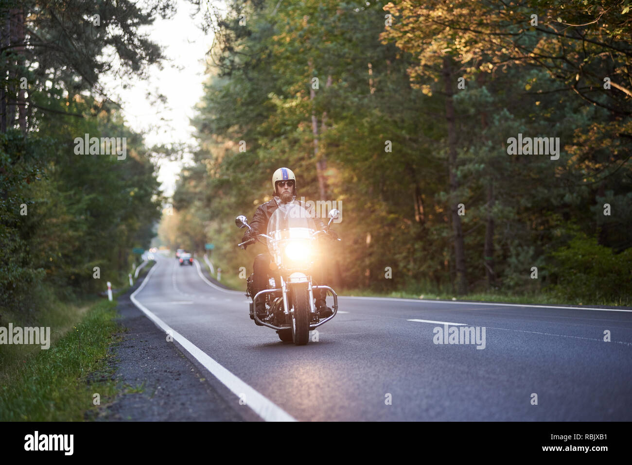 Barbuto biker in occhiali da sole, casco e pelle nera di equitazione abbigliamento moderni potenti cruiser motocicletta lungo la strada asfaltata che si snodano fra alti t verde Foto Stock