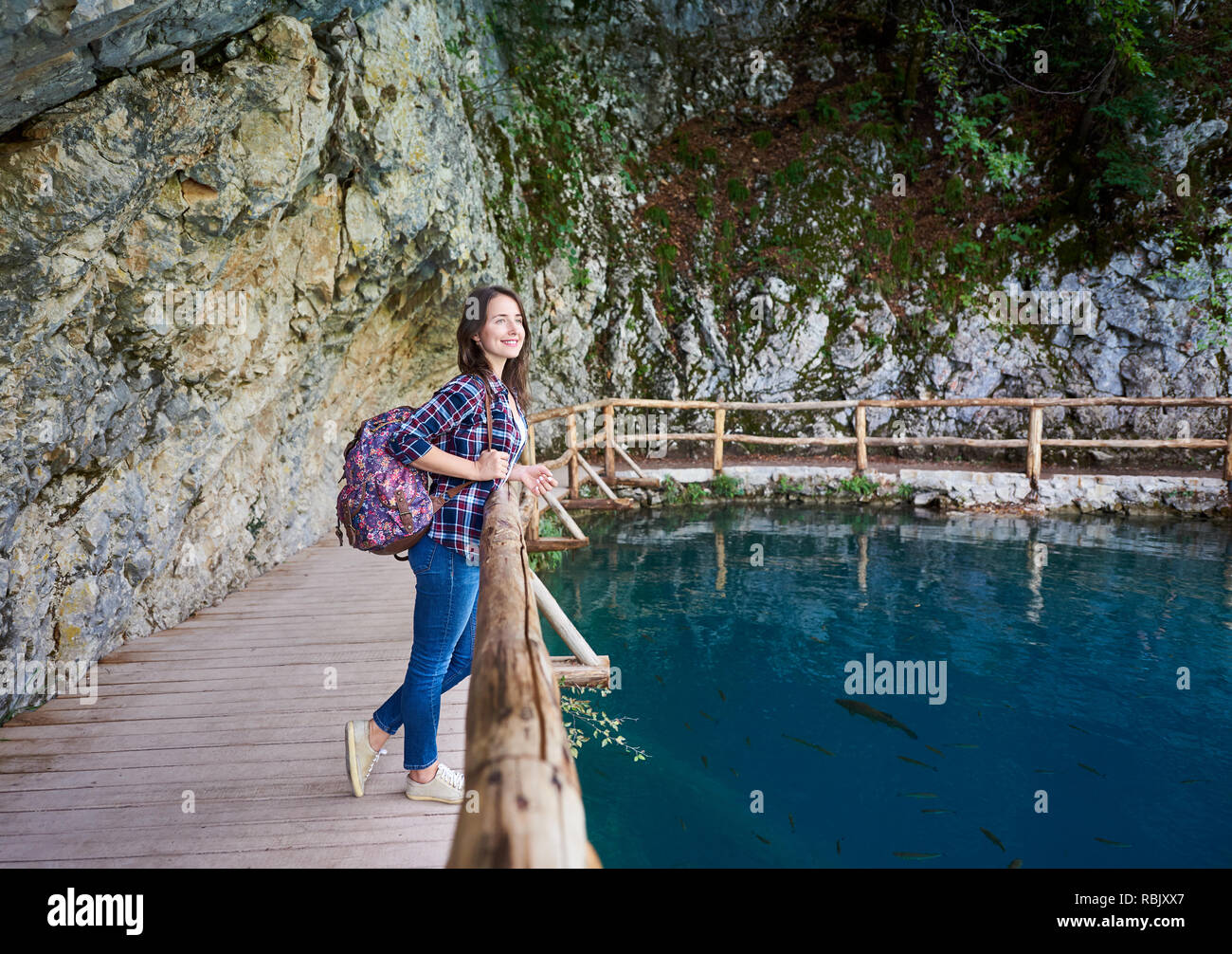 Slim piuttosto sorridente ragazza turistica con zaino in piedi sul percorso di legno sotto albero verde godendo di una splendida vista di pulito lago blu illuminato da estate su Foto Stock