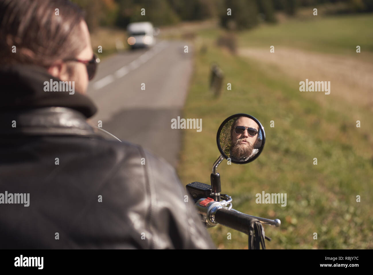 Bello barbuto biker in occhiali da sole scuri riflessa in moto mirror su sfondo sfocato di sunny road e verde paesaggio rurale. Foto Stock