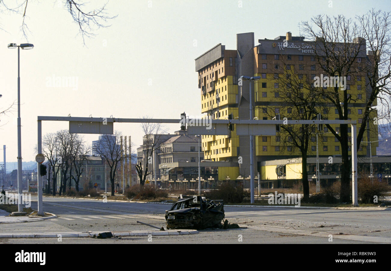 Il 15 marzo 1993 durante l'assedio di Sarajevo: un'auto bruciata sul vicolo di cecchino con l'Holiday Inn Hotel in background. Foto Stock