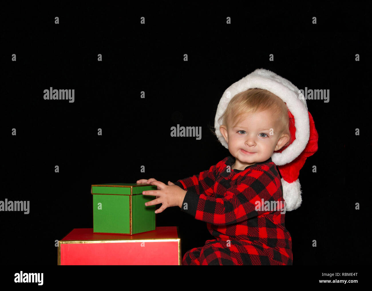 Close up ritratto di uno di un anno di età baby boy indossando un cappello da Babbo Natale a giocare con il rosso e il verde confezioni regalo guardando viewer con espressione comica. Bl Foto Stock