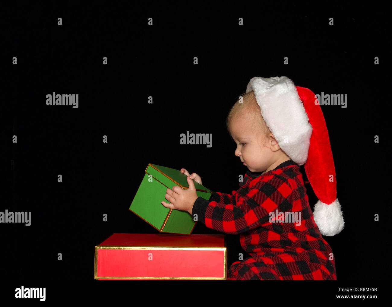 Close up ritratto di uno di un anno di età baby boy indossando un cappello da Babbo Natale a giocare con il rosso e il verde confezioni regalo, profilo visualizzare caselle di impilamento. Backgroun nero Foto Stock