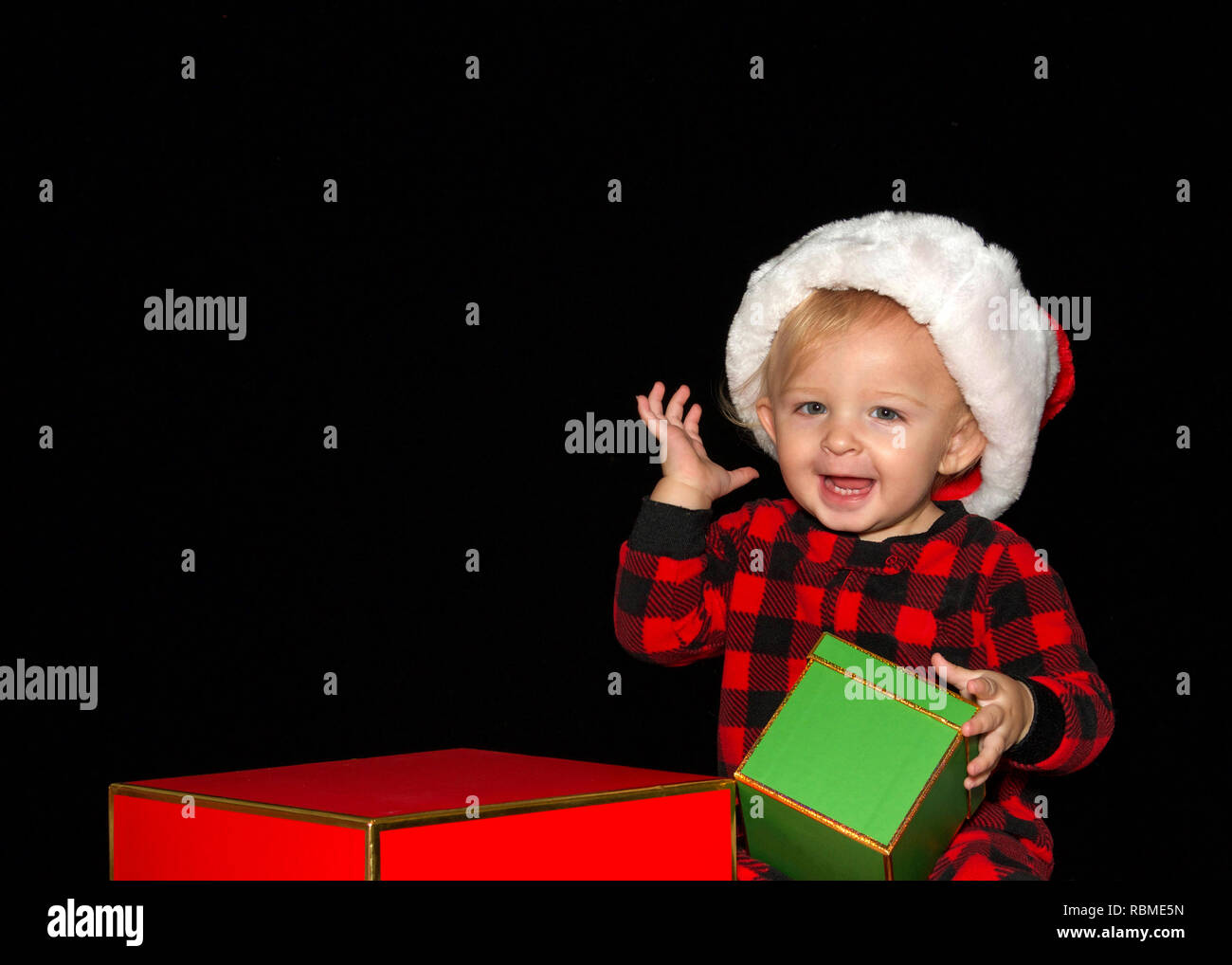 Close up ritratto di uno di un anno di età baby boy indossando un cappello da Babbo Natale a giocare con il rosso e il verde confezioni regalo, una mano grande sorriso con denti che mostra. Bl Foto Stock