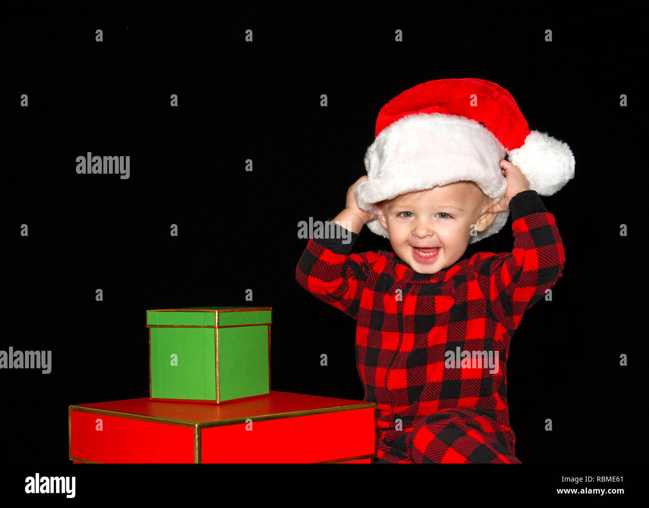 Close up ritratto di uno di un anno di età baby boy indossando un cappello da Babbo Natale a giocare con il rosso e il verde confezioni regalo, entrambe le mani sul cappello tirando sulla testa grande smil Foto Stock
