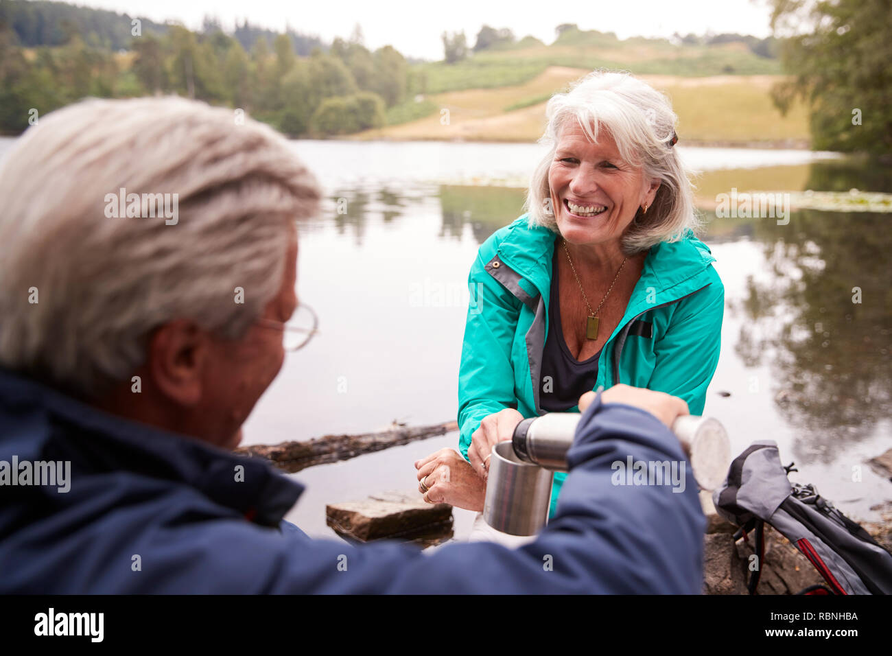 Coppia senior da un lago, uomo versando il caffè a sua moglie il cup, sulla spalla vista, Lake District, REGNO UNITO Foto Stock