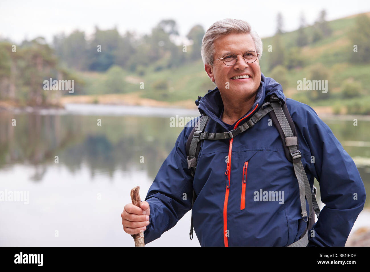 Uomo di mezza età in piedi da un lago tenendo un bastone sorridente, vicino Lake District, REGNO UNITO Foto Stock