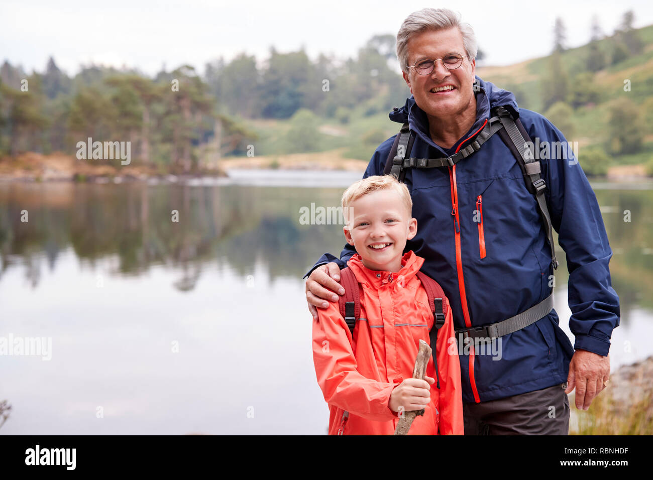 Nonno e nipote insieme in piedi sulla riva di un lago di sorridere alla telecamera, vicino Lake District, REGNO UNITO Foto Stock