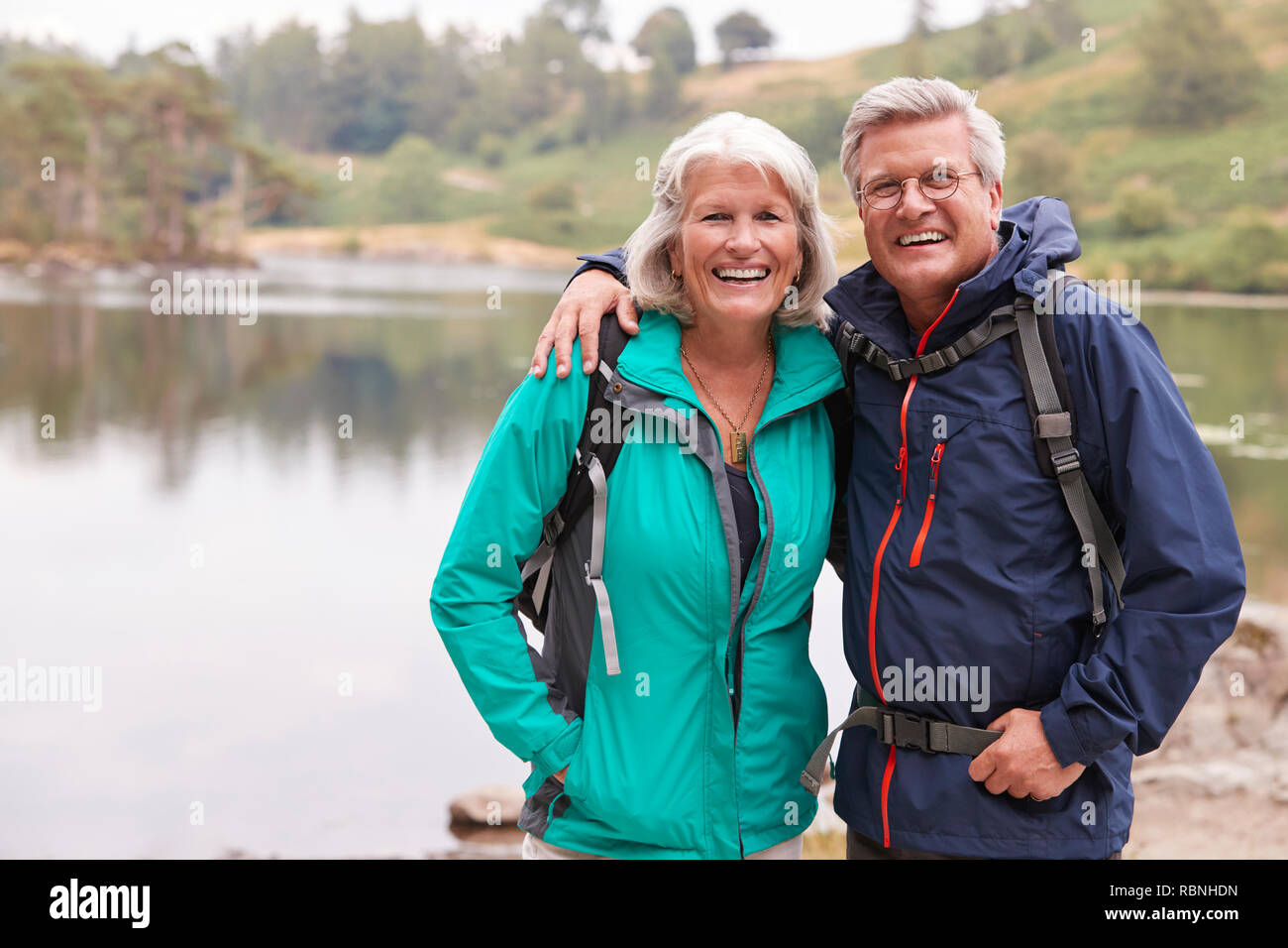 Felice coppia senior in piedi sulla riva di un lago di sorridere alla telecamera, Lake District, REGNO UNITO Foto Stock