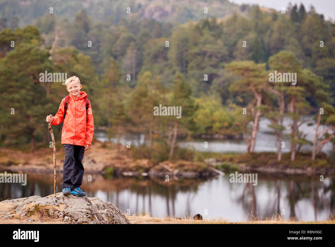 Un ragazzo in piedi su una roccia da un lago tenendo un bastone, sorridente alla telecamera, Lake District, REGNO UNITO Foto Stock