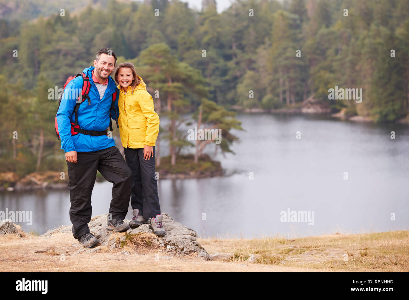 Caucasian pre-teen ragazza in piedi con suo padre su una roccia da un lago, sorridente alla telecamera, Lakeside sfondo Foto Stock