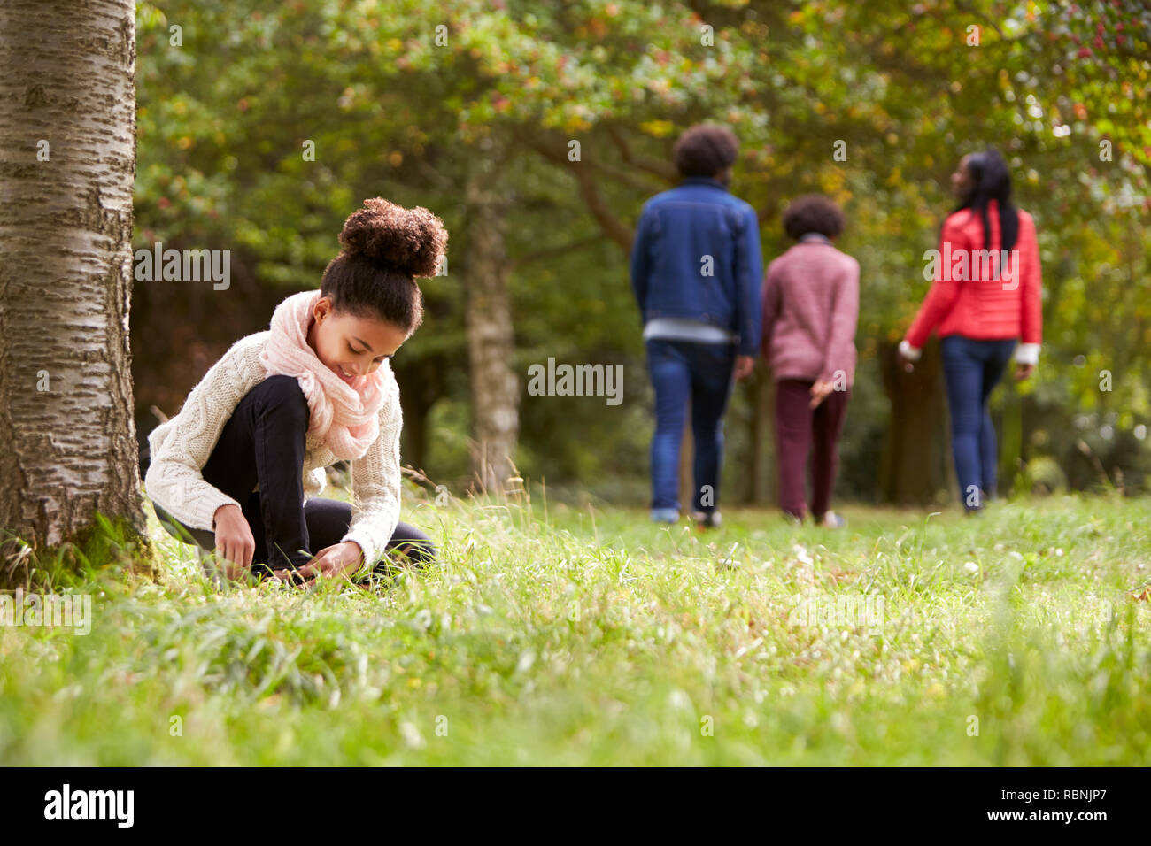 Razza mista ragazza inginocchiato nel parco di legare la sua scarpa, la sua famiglia passeggiate in background, angolo basso Foto Stock