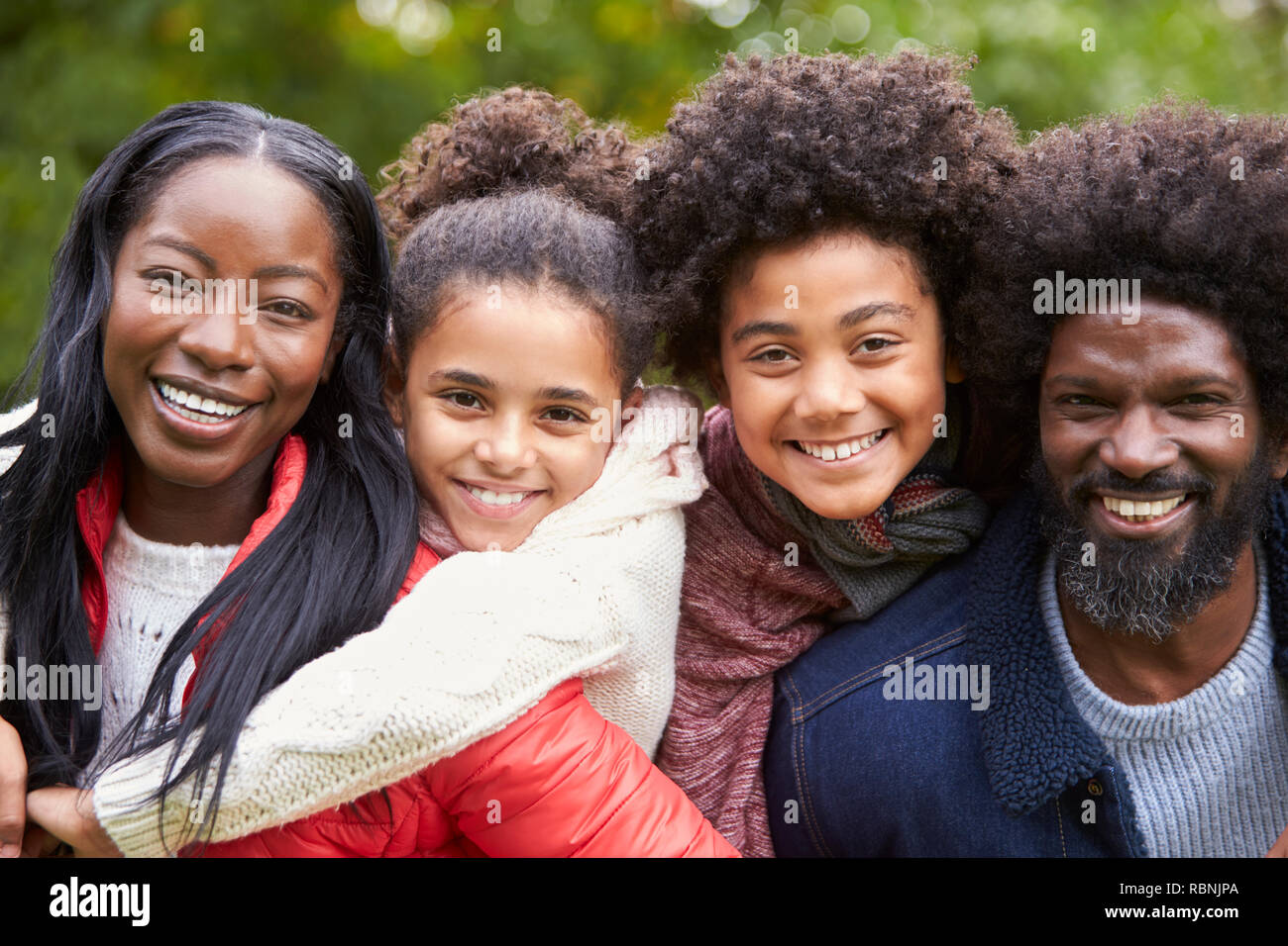 Razza mista famiglia sorridente alla telecamera, genitori piggybacking i bambini nel parco, vicino fino Foto Stock
