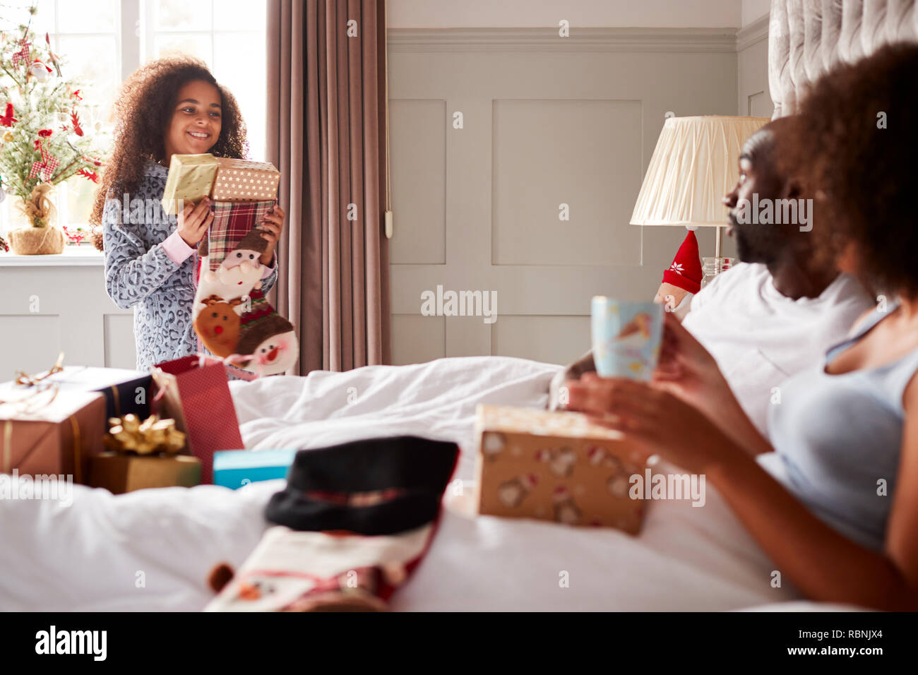 Giovani razza mista ragazza portando doni in suoi genitori di camera da letto la mattina di Natale, genitori seduta sul letto, close up Foto Stock
