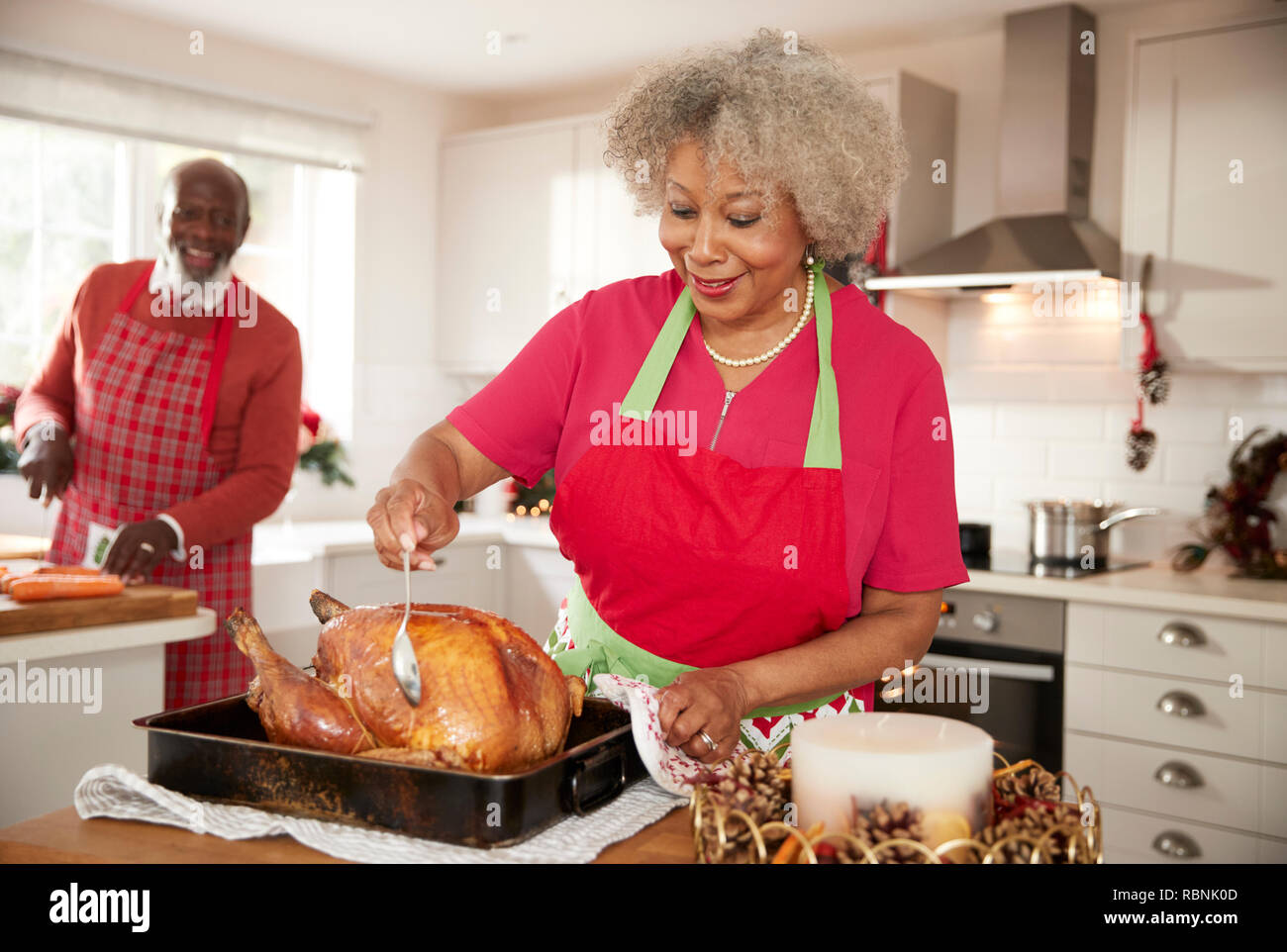 Senior nero donna inumidendo un tacchino arrosto in preparazione per la cena di Natale, suo marito tritare verdure in background, vista frontale, close up Foto Stock