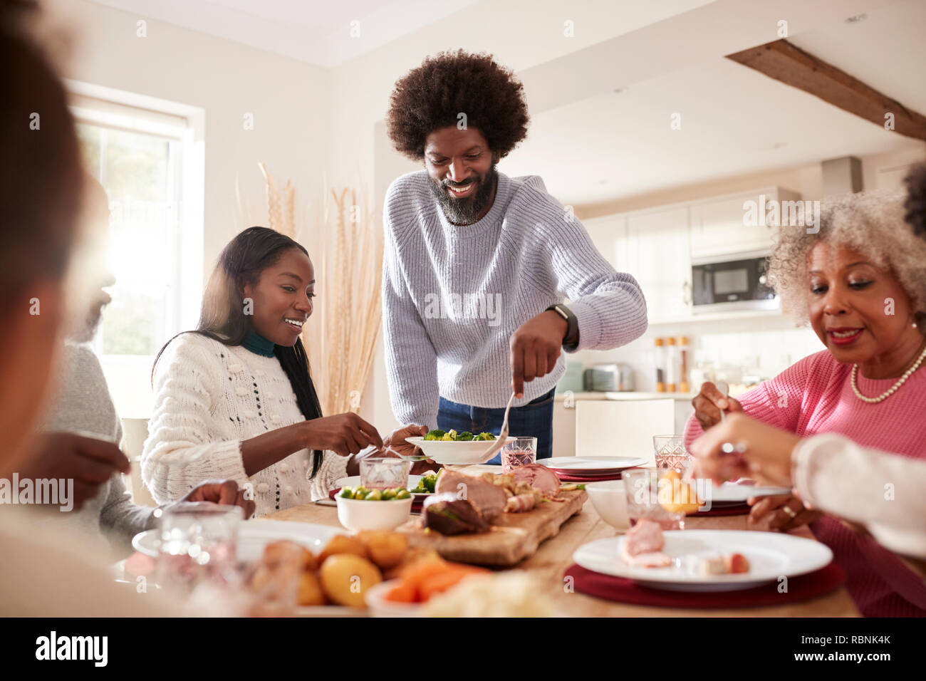 Persone di mezza età uomo nero carving e servire la carne a domenica cena in famiglia con il suo partner, i bambini e i loro nonni, vista frontale Foto Stock