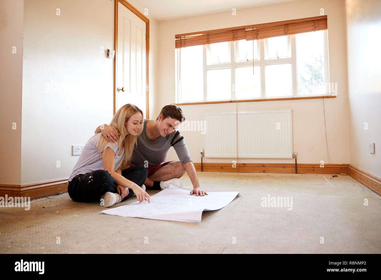 Giovane seduto sul pavimento guardando i piani in stanza vuota della nuova casa Foto Stock