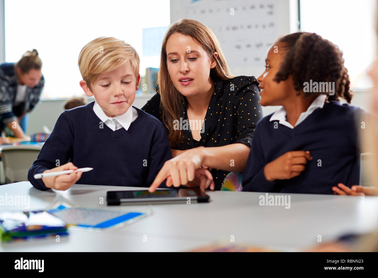 Femmina insegnante di scuola per aiutare due bambini usando un computer tablet al banco in una scuola primaria in aula, vista frontale Foto Stock