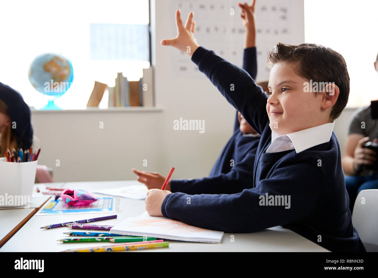 Scolaro con la sindrome di Down seduti ad una scrivania alzando la mano in una scuola primaria di classe, vicino, vista laterale Foto Stock