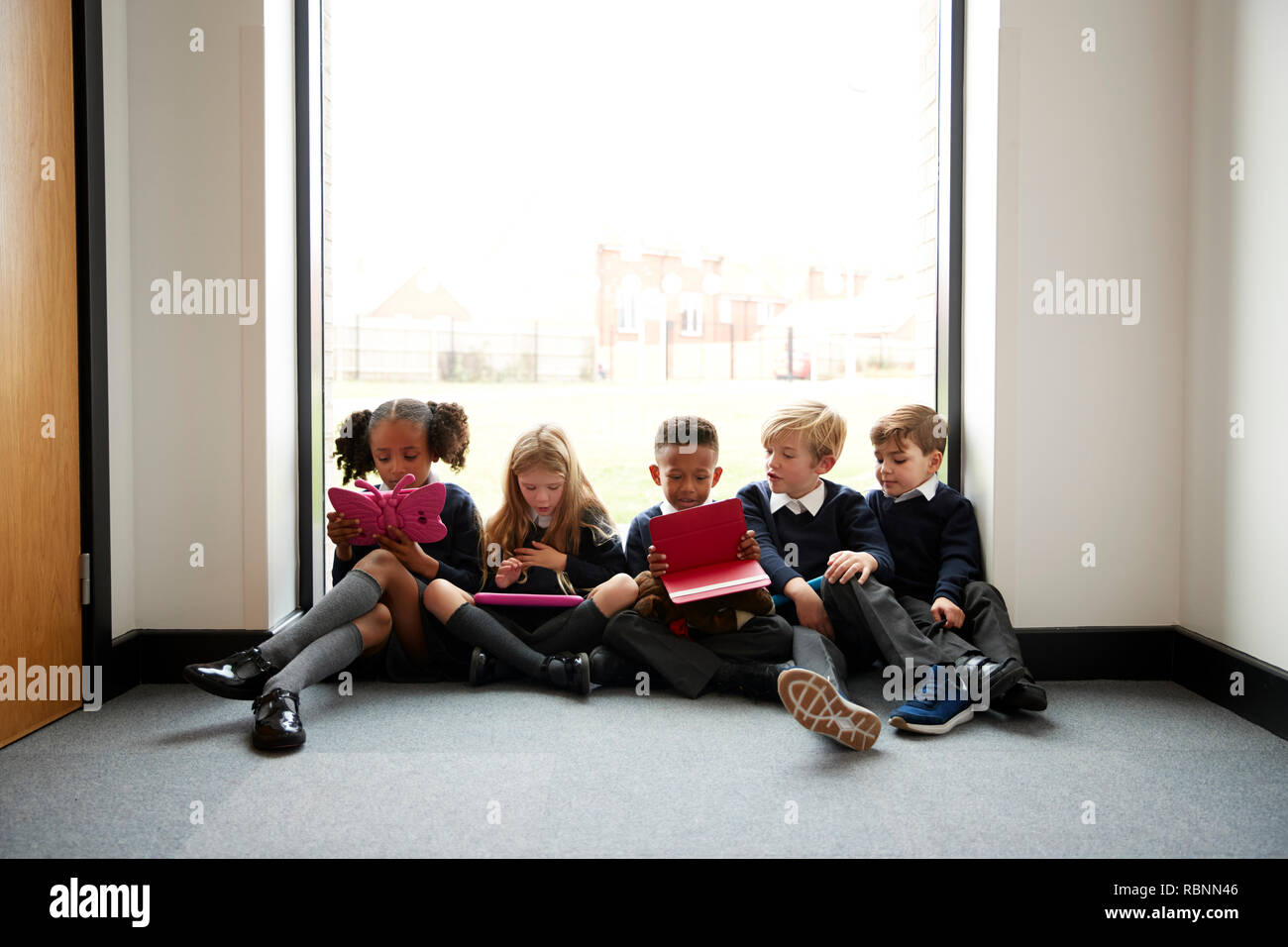 La scuola primaria i bambini seduti in una fila sul pavimento di fronte a una finestra in un corridoio di scuola utilizzando i computer tablet, vista frontale Foto Stock