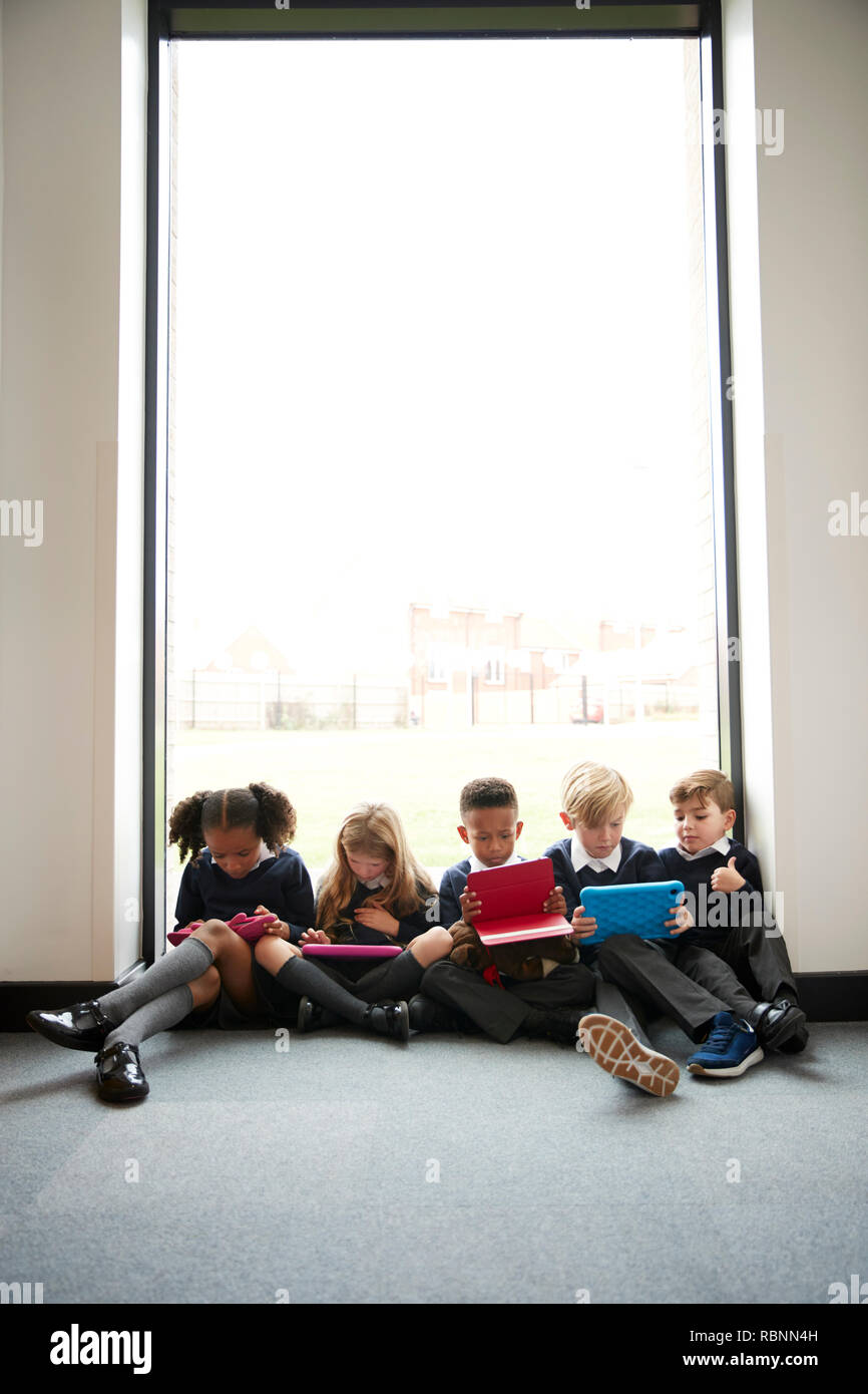 La scuola primaria i bambini seduti insieme sul pavimento di fronte a una finestra in un corridoio di scuola utilizzando i computer tablet, verticale Foto Stock