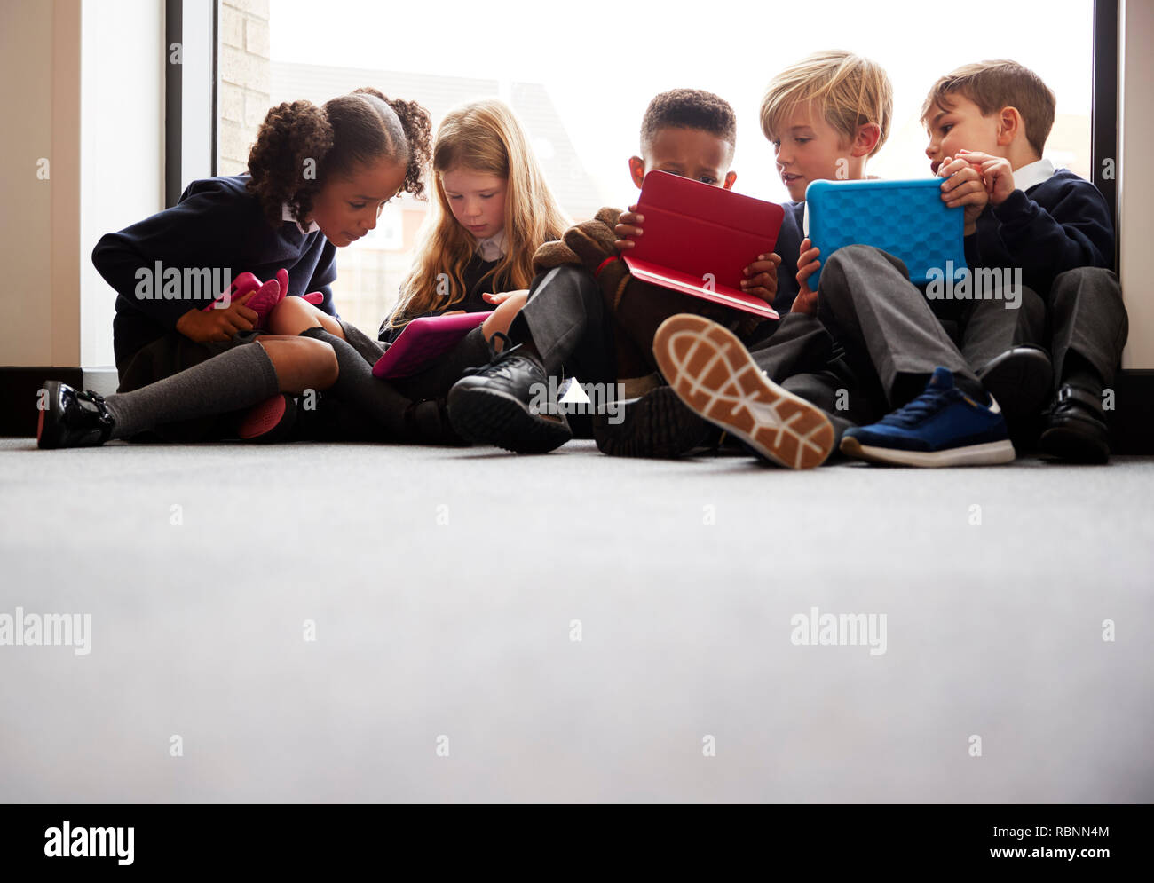 Basso angolo vista della scuola primaria amici seduti insieme davanti a una finestra in una scuola corridoio guardando i computer tablet insieme Foto Stock