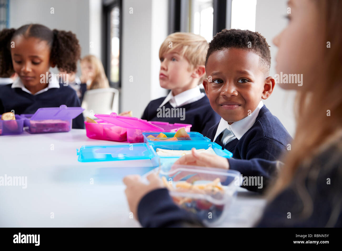 Sorridente scuola primaria i bambini seduti ad un tavolo a mangiare il loro pranzo al sacco insieme, il fuoco selettivo Foto Stock