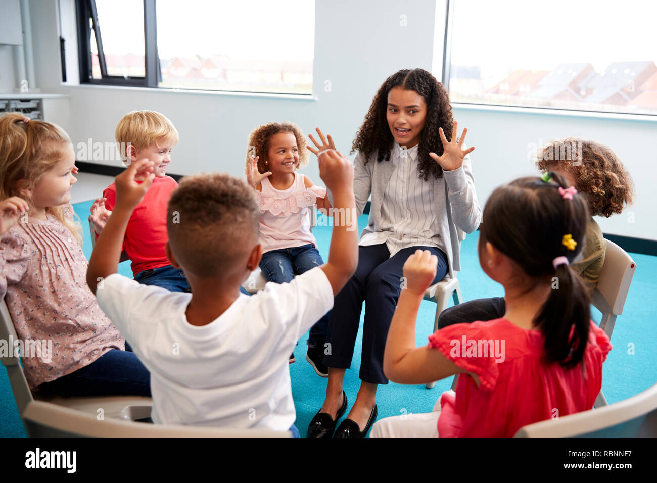 Una classe della scuola per neonati bambini seduti su sedie in un cerchio in aula, alzando le mani e imparare a contare con il loro insegnante femminile, close up Foto Stock