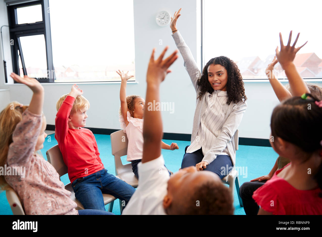 Una classe della scuola per neonati bambini seduti su sedie in un cerchio in aula, alzando le mani con il loro insegnante femminile, close up Foto Stock