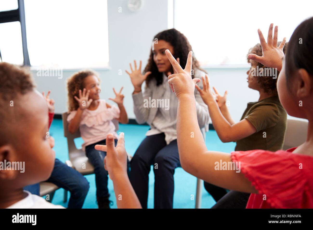 Una classe della scuola per neonati bambini seduti su sedie in un cerchio in aula, alzando le mani e imparare a contare con il loro insegnante femminile, close up Foto Stock