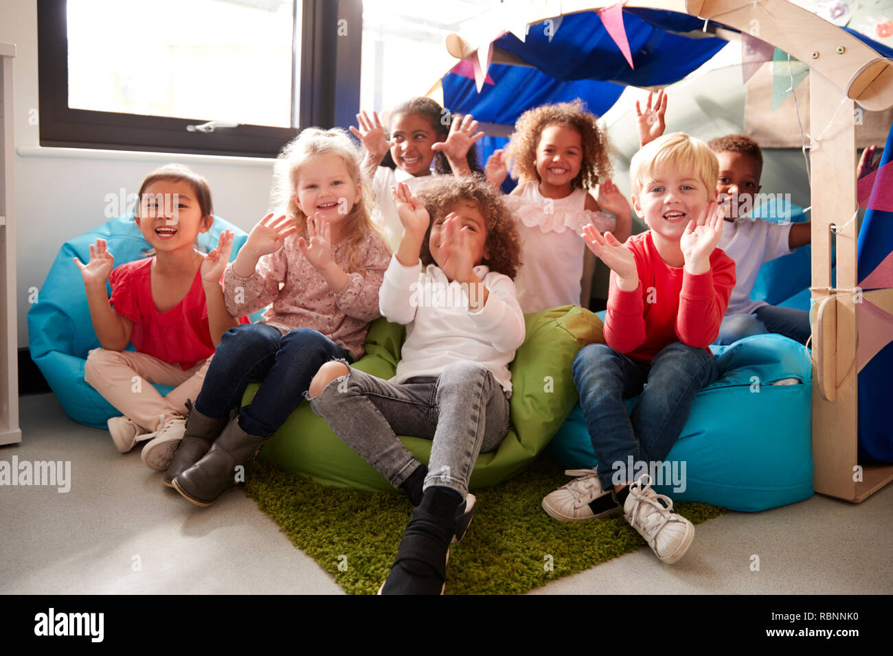 Un multi-etnico gruppo di scuole materne bambini seduti sui sacchi di fagioli in un confortevole angolo della classe, Sorridendo e agitando per fotocamera, angolo basso, close up Foto Stock